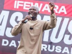 Salvation For All Souls Global Outreach Provides Free Medicals For Osun Indigenes, Begins Crusade April 27 Male speaker in a beige outfit on stage, microphone in hand, raising his right fist with a large event banner behind him.