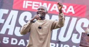 Salvation For All Souls Global Outreach Provides Free Medicals For Osun Indigenes, Begins Crusade April 27 Male speaker in a beige outfit on stage, microphone in hand, raising his right fist with a large event banner behind him.
