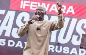 Salvation For All Souls Global Outreach Provides Free Medicals For Osun Indigenes, Begins Crusade April 27 Male speaker in a beige outfit on stage, microphone in hand, raising his right fist with a large event banner behind him.