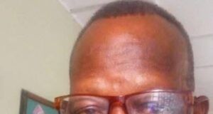 Renowned Veteran Journalist, Clement Ige, Dies At 68 Man with short hair and brown-framed glasses smiling at the camera indoors, wearing a red plaid shirt, with framed artwork in the background.