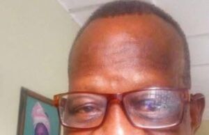 Renowned Veteran Journalist, Clement Ige, Dies At 68 Man with short hair and brown-framed glasses smiling at the camera indoors, wearing a red plaid shirt, with framed artwork in the background.