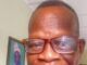 Renowned Veteran Journalist, Clement Ige, Dies At 68 Man with short hair and brown-framed glasses smiling at the camera indoors, wearing a red plaid shirt, with framed artwork in the background.