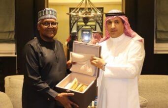 Saudi Envoy, Dangote Meet To Deepen Cooperation, Partnership Two men in traditional robes present an open box with gold-colored items inside, in a formal room.