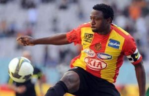 Ex-Super Eagles Striker Slumps, Dies During Training In Kaduna Soccer player in a red and yellow jersey kicking a ball during a match, with blurred stadium stands in the background.