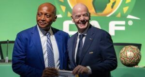 CAF Backs Infantino For Re-Election As FIFA President Two men in suits pose at a FIFA event, holding a FIFA banner; green backdrop with a display football nearby.