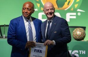CAF Backs Infantino For Re-Election As FIFA President Two men in suits pose at a FIFA event, holding a FIFA banner; green backdrop with a display football nearby.