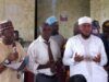 2026 Hajj: Oyo Muslim Pilgrims’ Welfare Board Concludes Vaccination Exercise Group of men at a community meeting; one man speaks into a microphone while two others stand nearby, with a crowd seated in the background.