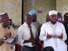 2026 Hajj: Oyo Muslim Pilgrims’ Welfare Board Concludes Vaccination Exercise Group of men at a community meeting; one man speaks into a microphone while two others stand nearby, with a crowd seated in the background.