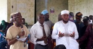 2026 Hajj: Oyo Muslim Pilgrims’ Welfare Board Concludes Vaccination Exercise Group of men at a community meeting; one man speaks into a microphone while two others stand nearby, with a crowd seated in the background.