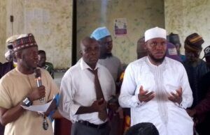 2026 Hajj: Oyo Muslim Pilgrims’ Welfare Board Concludes Vaccination Exercise Group of men at a community meeting; one man speaks into a microphone while two others stand nearby, with a crowd seated in the background.