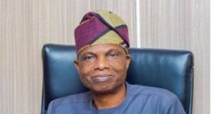 Oyo Govt. Launches Free Evening Market Classes To Tackle Out-Of-School Menace Man wearing a purple‑green headwrap and blue traditional outfit, seated in a dark leather chair.