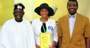 We’ll Not Remove Tinubu’s Wife As Pastor In RCCG – Adeboye Three people pose indoors: a smiling man in white traditional attire, a woman in a white jacket and black hat holding a yellow certificate, and a man in a brown suit.