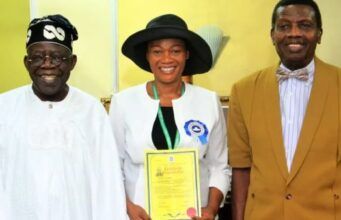 We’ll Not Remove Tinubu’s Wife As Pastor In RCCG – Adeboye Three people pose indoors: a smiling man in white traditional attire, a woman in a white jacket and black hat holding a yellow certificate, and a man in a brown suit.