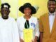 We’ll Not Remove Tinubu’s Wife As Pastor In RCCG – Adeboye Three people pose indoors: a smiling man in white traditional attire, a woman in a white jacket and black hat holding a yellow certificate, and a man in a brown suit.