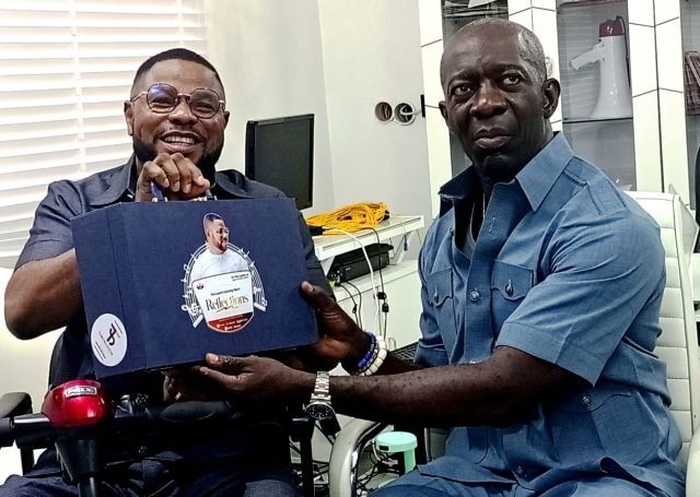 Adekanmbi with Ayefele Two men share a moment in a bright office: one in a wheelchair smiles while handing a blue gift box to a man in a blue shirt who sits nearby. The box has a circular sticker on its front.