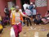 Eid-L-Kabir: OYSG Aids Workers With Subsidized Commodities Aid workers in orange vests distribute supplies to people outside a red building; a yellow vehicle is parked on the left while residents wait nearby.