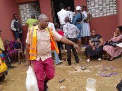Eid-L-Kabir: OYSG Aids Workers With Subsidized Commodities Aid workers in orange vests distribute supplies to people outside a red building; a yellow vehicle is parked on the left while residents wait nearby.