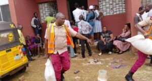 Eid-L-Kabir: OYSG Aids Workers With Subsidized Commodities Aid workers in orange vests distribute supplies to people outside a red building; a yellow vehicle is parked on the left while residents wait nearby.