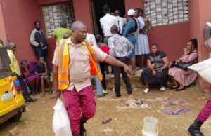 Eid-L-Kabir: OYSG Aids Workers With Subsidized Commodities Aid workers in orange vests distribute supplies to people outside a red building; a yellow vehicle is parked on the left while residents wait nearby.