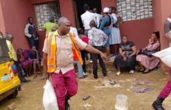 Eid-L-Kabir: OYSG Aids Workers With Subsidized Commodities Aid workers in orange vests distribute supplies to people outside a red building; a yellow vehicle is parked on the left while residents wait nearby.