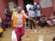 Eid-L-Kabir: OYSG Aids Workers With Subsidized Commodities Aid workers in orange vests distribute supplies to people outside a red building; a yellow vehicle is parked on the left while residents wait nearby.