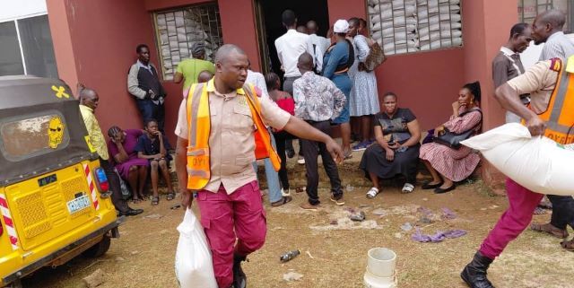 Aids For Oyo Aid workers in orange vests distribute supplies to people outside a red building; a yellow vehicle is parked on the left while residents wait nearby.