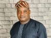Senator Alli mourns ex-Egbeda LG boss, Akeem Sadiq Man in a dark pinstriped tunic with white piping and a brown-orange striped headwrap, standing in front of a light gray stone wall.