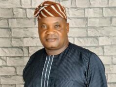 Senator Alli Mourns Ex-Egbeda LG Boss, Akeem Sadiq Man in a dark pinstriped tunic with white piping and a brown-orange striped headwrap, standing in front of a light gray stone wall.