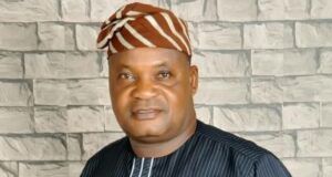 Senator Alli Mourns Ex-Egbeda LG Boss, Akeem Sadiq Man in a dark pinstriped tunic with white piping and a brown-orange striped headwrap, standing in front of a light gray stone wall.