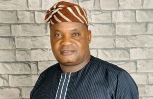 Senator Alli Mourns Ex-Egbeda LG Boss, Akeem Sadiq Man in a dark pinstriped tunic with white piping and a brown-orange striped headwrap, standing in front of a light gray stone wall.