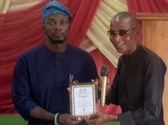 Oyo NUJ Chairman, Abas Establishes Prizes For Poly Ibadan Mass Communication Students Two men on a stage exchange an award plaque; the man on the left in blue traditional attire presents it to the man on the right, who holds a microphone and smiles.