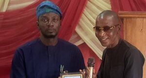Oyo NUJ Chairman, Abas Establishes Prizes For Poly Ibadan Mass Communication Students Two men on a stage exchange an award plaque; the man on the left in blue traditional attire presents it to the man on the right, who holds a microphone and smiles.