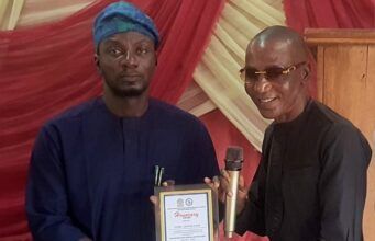 Oyo NUJ Chairman, Abas Establishes Prizes For Poly Ibadan Mass Communication Students Two men on a stage exchange an award plaque; the man on the left in blue traditional attire presents it to the man on the right, who holds a microphone and smiles.