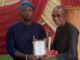 Oyo NUJ Chairman, Abas Establishes Prizes For Poly Ibadan Mass Communication Students Two men on a stage exchange an award plaque; the man on the left in blue traditional attire presents it to the man on the right, who holds a microphone and smiles.