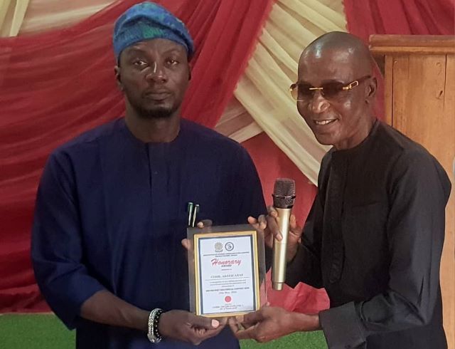 Akeen Abas Two men on a stage exchange an award plaque; the man on the left in blue traditional attire presents it to the man on the right, who holds a microphone and smiles.