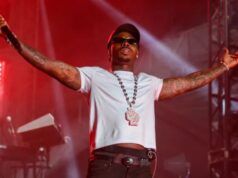 Asake Announces ‘In God We Trust’ Tour With Uncle Waffles Male rapper on stage with arms outstretched, wearing a white crop tee, cap, sunglasses, and a large pendant necklace under red stage lights.