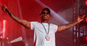 Asake Announces ‘In God We Trust’ Tour With Uncle Waffles Male rapper on stage with arms outstretched, wearing a white crop tee, cap, sunglasses, and a large pendant necklace under red stage lights.