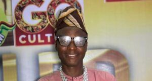 August24news.com Publisher, Ajagbe, Bags Media Personality Award Smiling man in a pink traditional outfit and head wrap holding a gold star trophy on stage.