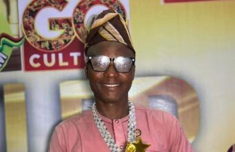 August24news.com Publisher, Ajagbe, Bags Media Personality Award Smiling man in a pink traditional outfit and head wrap holding a gold star trophy on stage.