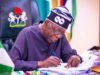 Tinubu’s Govt To Scrap JSS/SSS Structure Over 20m Dropouts Nigerian official signing documents at a desk, with the coat of arms on the chair behind him.