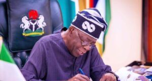 Tinubu’s Govt To Scrap JSS/SSS Structure Over 20m Dropouts Nigerian official signing documents at a desk, with the coat of arms on the chair behind him.