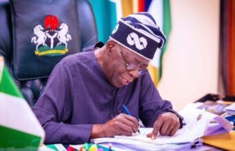 Tinubu’s Govt Announces Seven-Week Roadmap For National Policing Bill, Invites Public Input Nigerian official signing documents at a desk, with the coat of arms on the chair behind him.