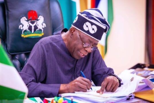 Tinubu’s Govt Awards Contracts To Rehabilitate 13 Police Training Institutions Nigerian official signing documents at a desk, with the coat of arms on the chair behind him.
