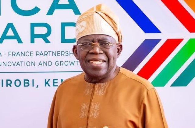 Bola Tinubu Smiling man in tan traditional attire and head wrap posing in front of an Africa-France partnership banner at a formal event