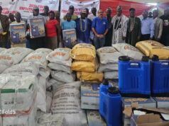 Excitement As Dangote Cement Gboko Rolls Out Multi-Million Naira Empowerment Programmes For Six Host Communities In Benue Group of people standing behind large piles of sacks and blue jerrycans at a donation event, with banners in the background.