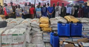 Excitement As Dangote Cement Gboko Rolls Out Multi-Million Naira Empowerment Programmes For Six Host Communities In Benue Group of people standing behind large piles of sacks and blue jerrycans at a donation event, with banners in the background.