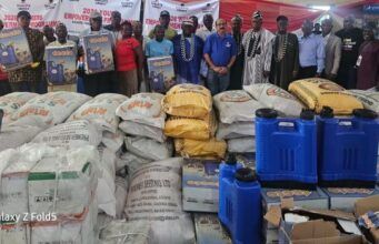 Excitement As Dangote Cement Gboko Rolls Out Multi-Million Naira Empowerment Programmes For Six Host Communities In Benue Group of people standing behind large piles of sacks and blue jerrycans at a donation event, with banners in the background.
