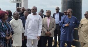 Dangote Hosts Femi Otedola, First HoldCo Executives At Refinery Group of formally dressed people standing on a pier by a railing, listening to a speaker near the water.