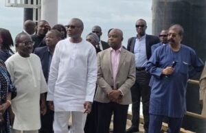 Dangote Hosts Femi Otedola, First HoldCo Executives At Refinery Group of formally dressed people standing on a pier by a railing, listening to a speaker near the water.