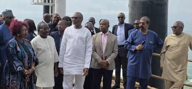 Dangote-Otedola Group of formally dressed people standing on a pier by a railing, listening to a speaker near the water.